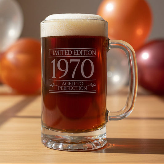 A clear glass beer stein mug filled with dark amber beer, topped with white foam, standing on a light wood surface against a background of balloons. The mug is etched with the text 'LIMITED EDITION 1970 AGED TO PERFECTION'.