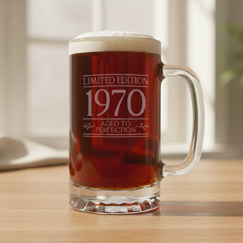 A clear glass beer stein mug filled with dark amber beer, topped with white foam, standing on a light wood surface against a neutral  background. The mug is etched with the text 'LIMITED EDITION 1970 AGED TO PERFECTION'.