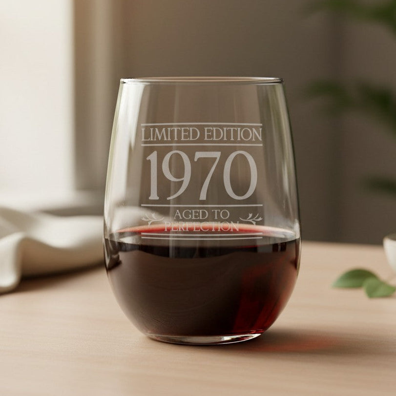 A clear stemless wine glass with red wine inside, engraved with 'LIMITED EDITION 1970 AGED TO PERFECTION', set on a wooden table in a softly lit room.