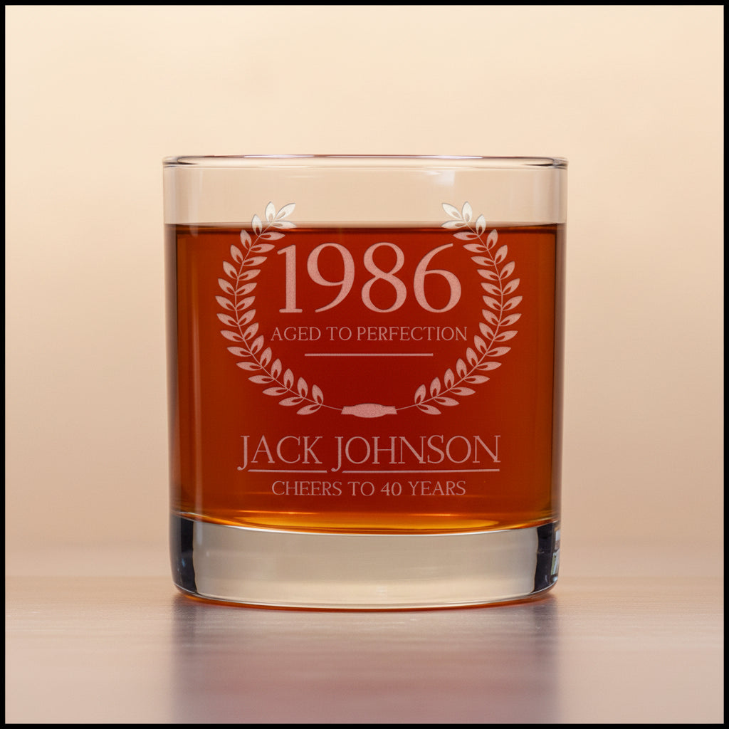 Glass tumbler with engraved text '1986 Aged to Perfection, Jack Johnson Cheers to 40 Years' on a light beige background.