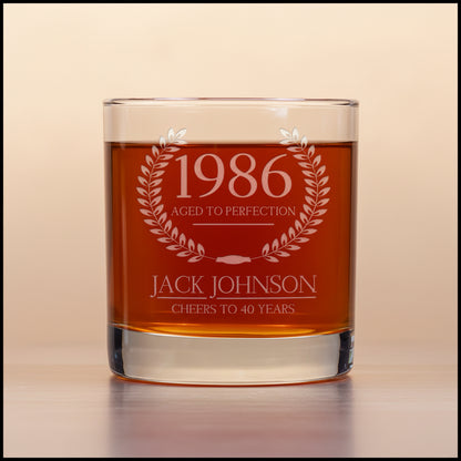 Glass tumbler with engraved text '1986 Aged to Perfection, Jack Johnson Cheers to 40 Years' on a light beige background.