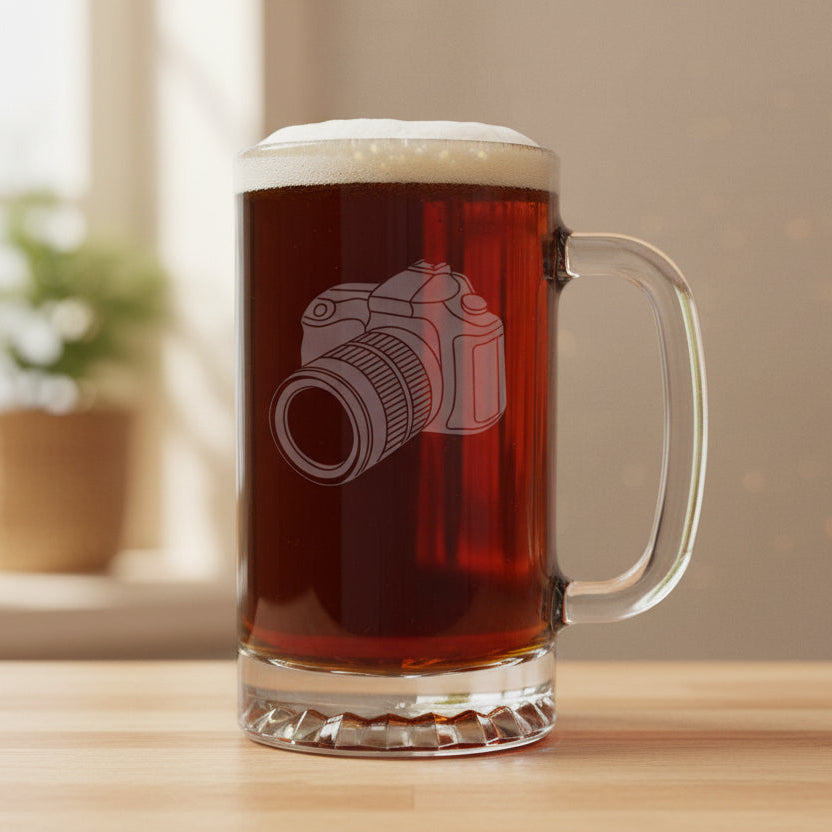 A clear glass beer mug filled with dark beer, featuring an etched camera design on the front, set on a light wood surface.