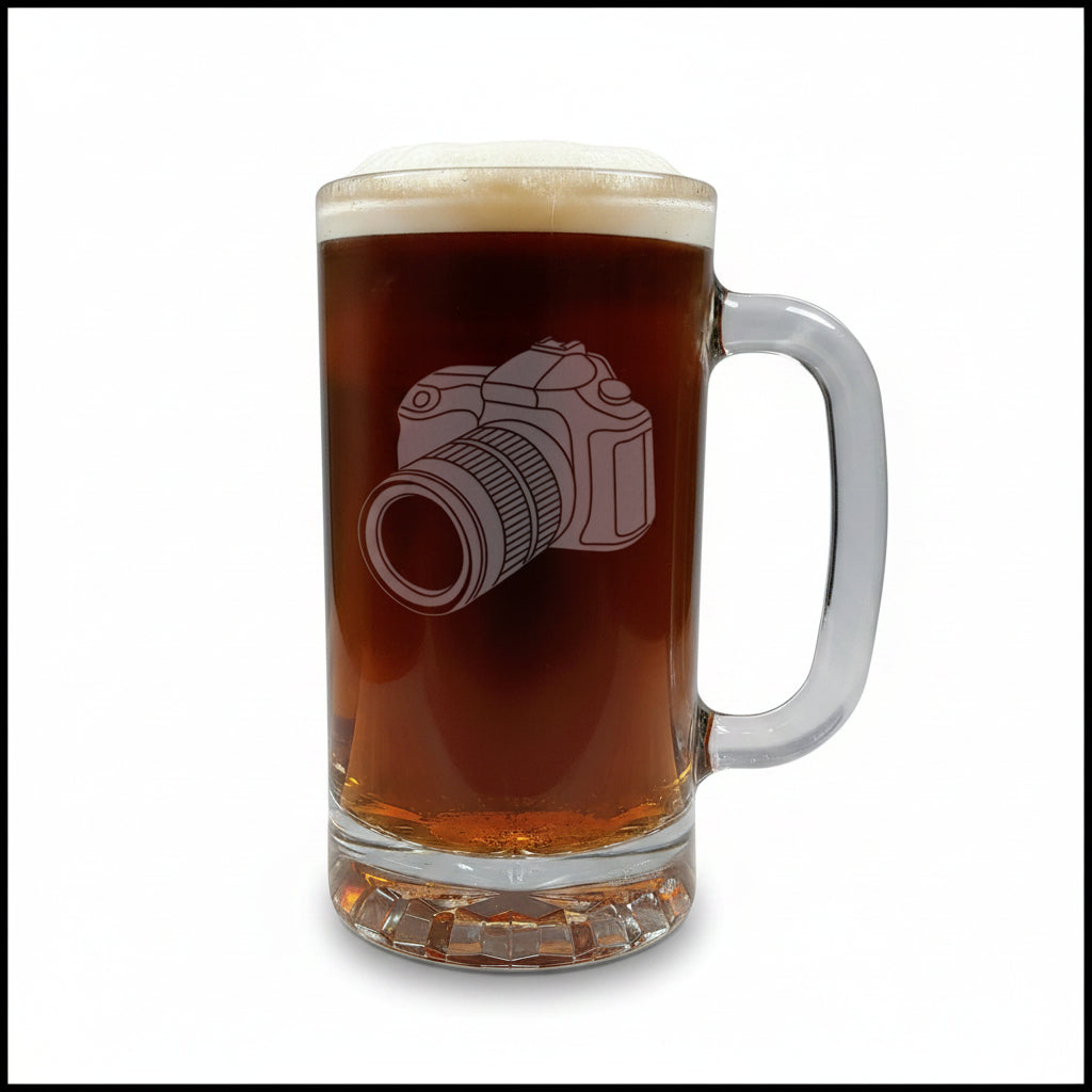 A clear glass beer mug filled with dark beer, featuring an etched camera graphic on the front, set on a light wood surface against a white background.