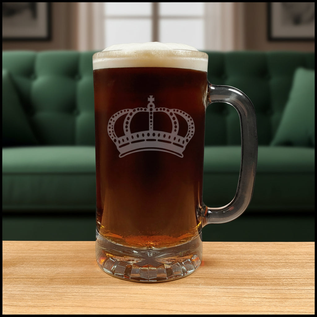 A clear glass beer mug with a handle, filled with dark amber beer and topped with white foam, featuring a white crown design  on the front, set on a light wood surface with a green couch  background. 
