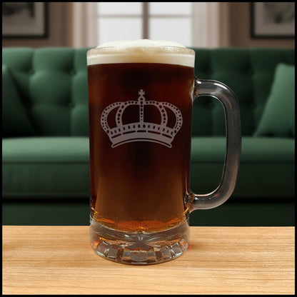 A clear glass beer mug with a handle, filled with dark amber beer and topped with white foam, featuring a white crown design  on the front, set on a light wood surface with a green couch  background. 