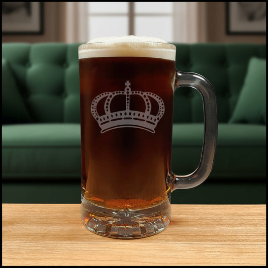 A clear glass beer mug with a handle, filled with dark amber beer and topped with white foam, featuring a white crown design  on the front, set on a light wood surface with a green couch  background. 
