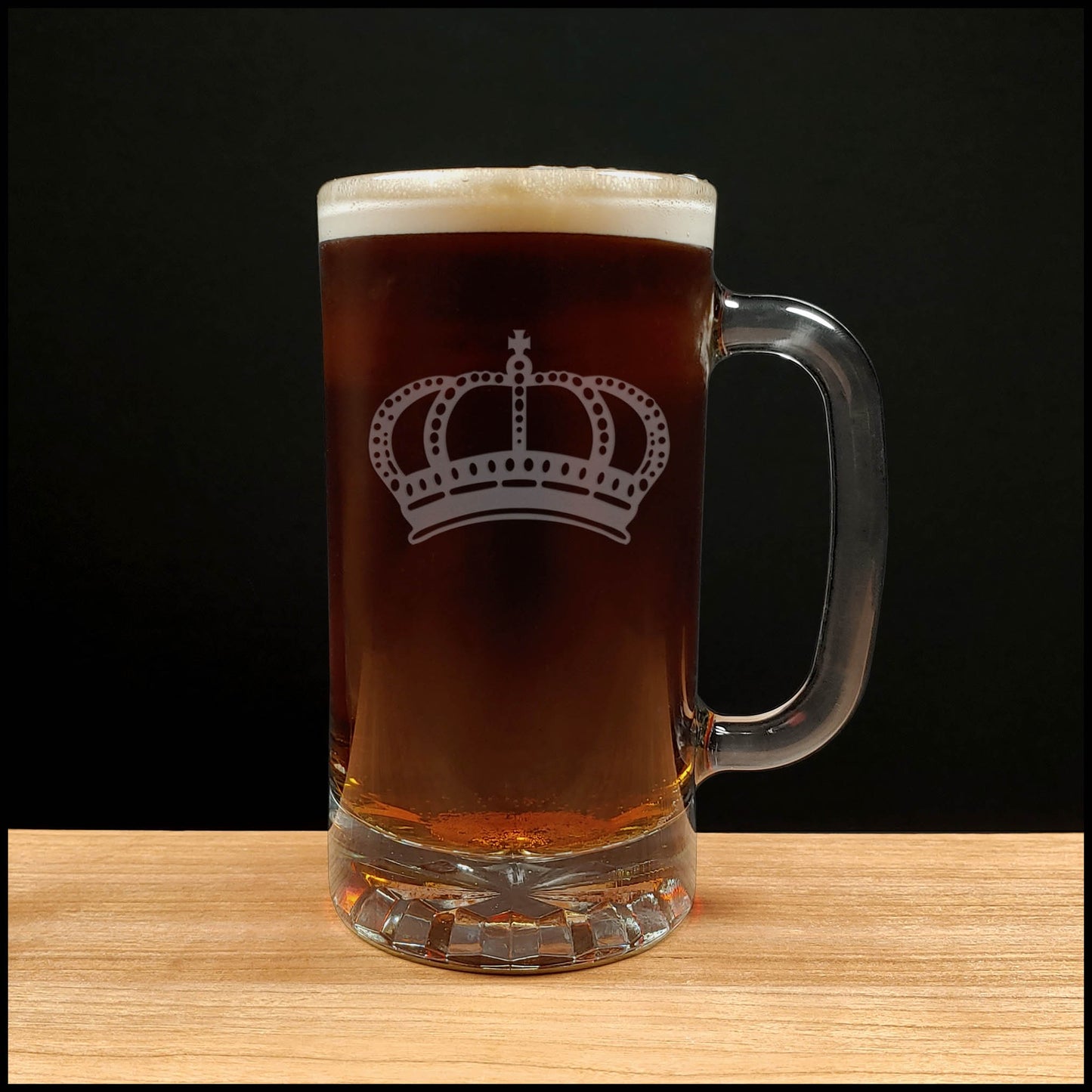 A clear glass beer mug with a handle, filled with dark amber beer and topped with white foam, featuring a white crown graphic on the front, set on a light wood surface against a black background.
