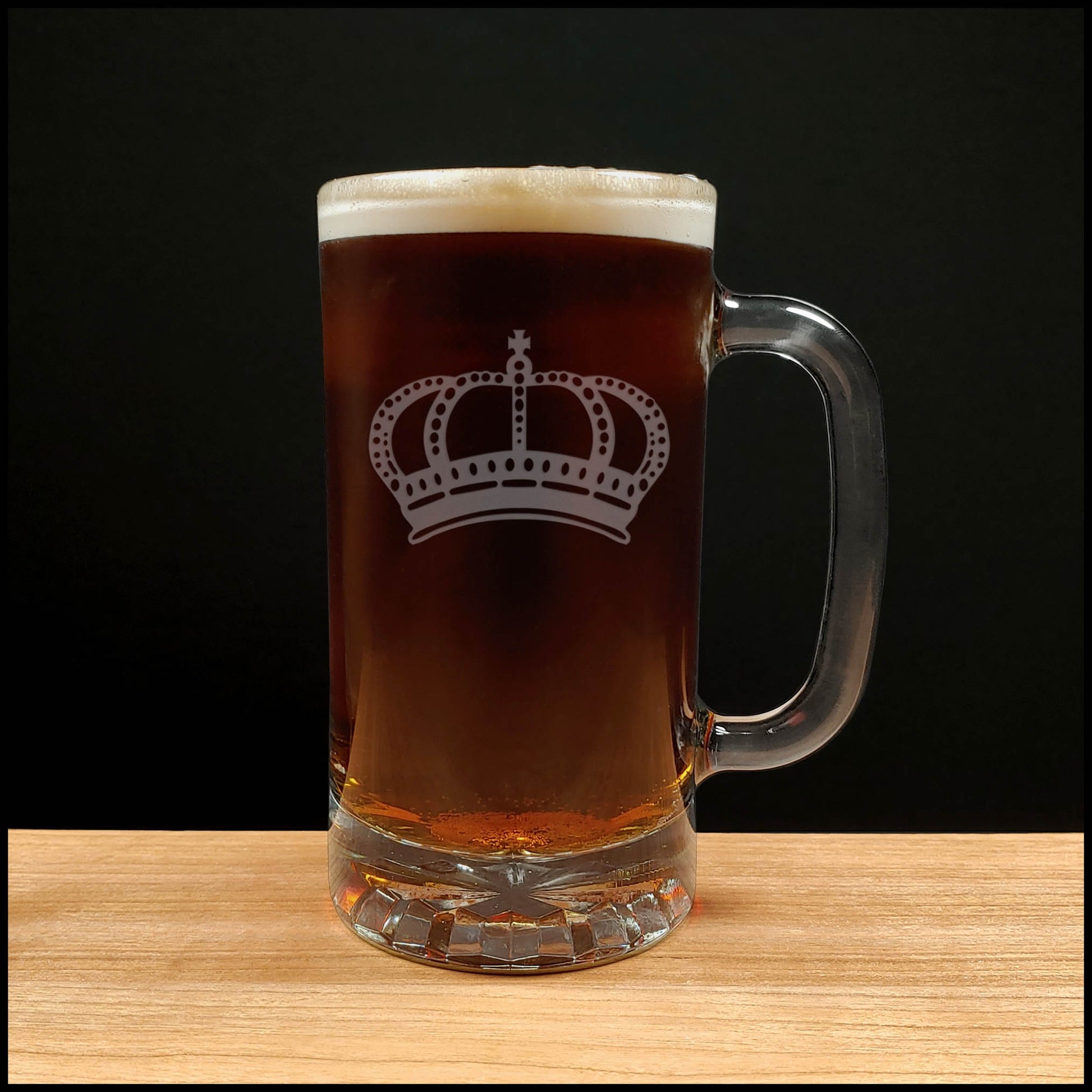 A clear glass beer mug with a handle, filled with dark amber beer and topped with white foam, featuring a white crown graphic on the front, set on a light wood surface against a black background.