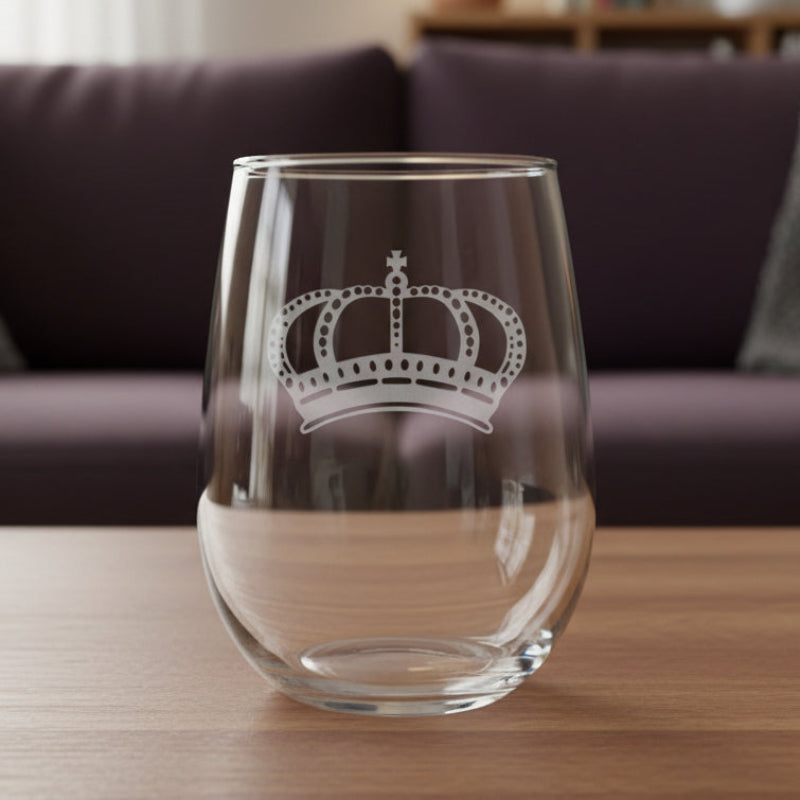 A clear stemless wine glass with an etched silver crown design, resting on a wooden surface with a purple couch in the background.