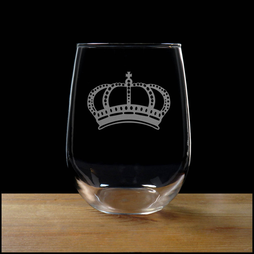 A clear stemless wine glass with an etched silver crown design, resting on a wooden surface against a solid black background.