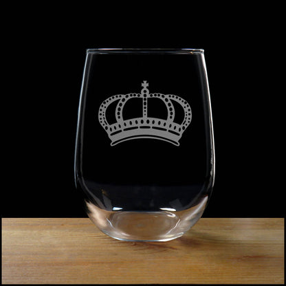 A clear stemless wine glass with an etched silver crown design, resting on a wooden surface against a solid black background.