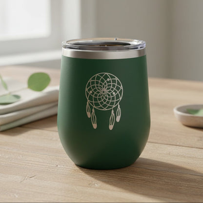 A dark green insulated stemless tumbler with a clear lid and a white dreamcatcher design on the front, set on a light wooden table.
