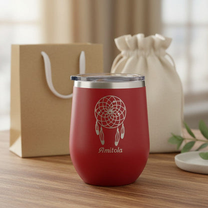 A red insulated stemless wine tumbler with a clear lid, featuring a white dreamcatcher design and the name 'Amitola' printed on the front. The tumbler is set on a wooden table with a kraft paper gift bag and a cream drawstring pouch in a softly blurred background.