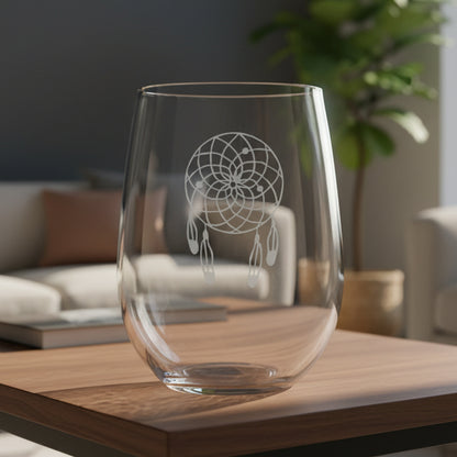 A clear stemless wine glass with an etched dreamcatcher design, resting on a wooden surface with a living room in the background.