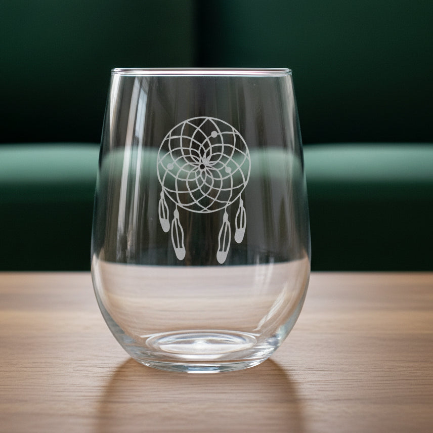 A clear stemless wine glass with an etched dreamcatcher design, resting on a wooden surface with a green couch in the background.