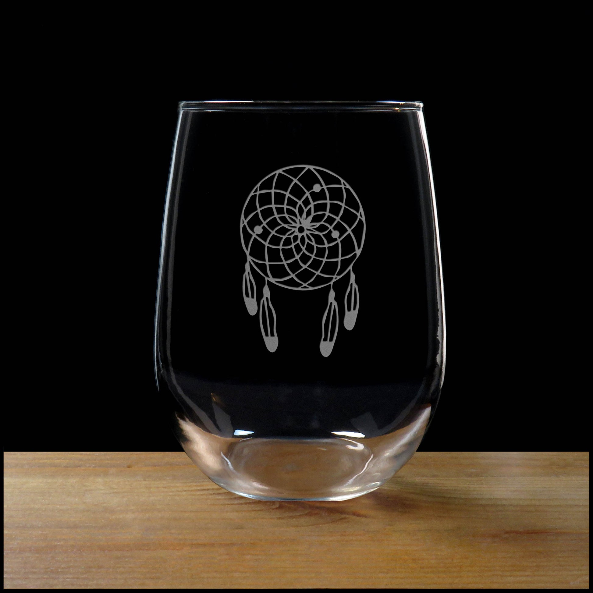 A clear stemless wine glass with an etched dreamcatcher design, resting on a wooden surface against a solid black background.