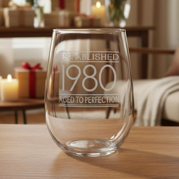 Clear glass with engraved text 'Established 1980 Aged to Perfection' on a wooden surface 