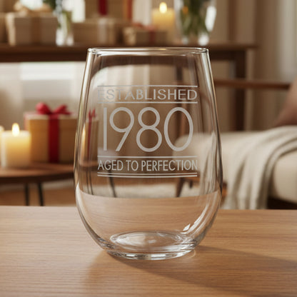 Clear glass with engraved text 'Established 1980 Aged to Perfection' on a wooden surface 