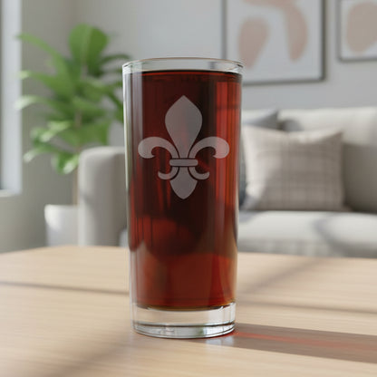 A clear pint glass filled with dark beer, featuring a white fleur-de-lis emblem on the front, set against a lving room.
