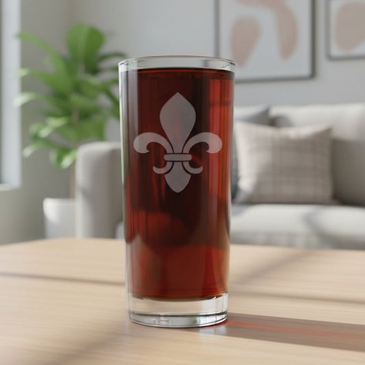A clear pint glass filled with dark beer, featuring a white fleur-de-lis emblem on the front, set against a lving room.