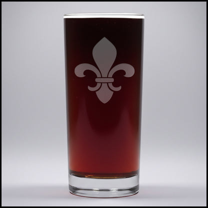 A clear pint glass filled with dark beer, featuring a white fleur-de-lis emblem on the front, set against a little gray background.