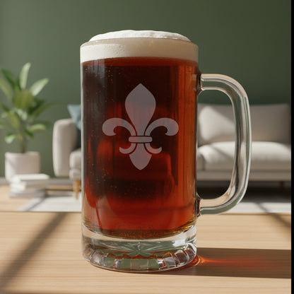 A clear glass beer mug with a handle, filled with dark brown beer and topped with white foam, featuring a white fleur-de-lis symbol on the front. The mug is placed on a light wood surface against a living room background.