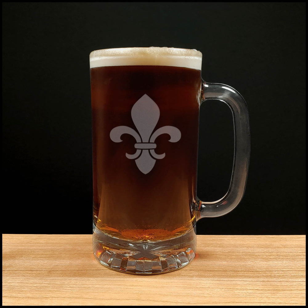 A clear glass beer mug with a handle, filled with dark brown beer and topped with white foam, featuring a white fleur-de-lis symbol on the front. The mug is placed on a light wood surface against a solid black background.