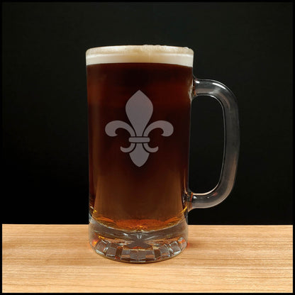 A clear glass beer mug with a handle, filled with dark brown beer and topped with white foam, featuring a white fleur-de-lis symbol on the front. The mug is placed on a light wood surface against a solid black background.