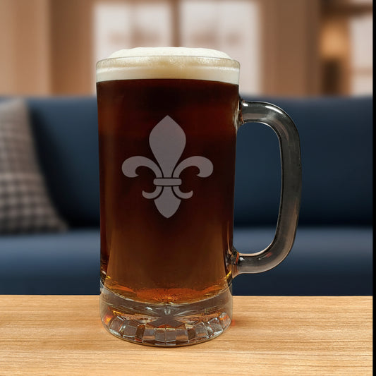 A clear glass beer mug with a handle, filled with dark brown beer and topped with white foam, featuring a white fleur-de-lis symbol on the front. The mug is placed on a light wood surface 