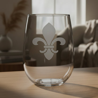 A clear stemless wine glass with a gray fleur-de-lis symbol etched on the front, resting on a wooden surface 