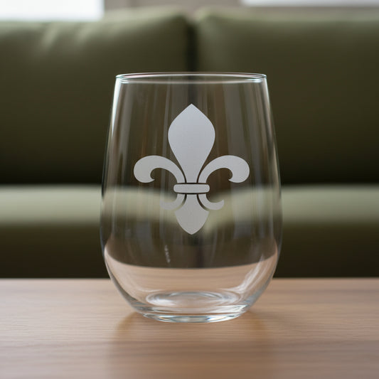 A clear stemless wine glass fleur-de-lis symbol etched on the front, resting on a wooden surface with a green couch in the background.
