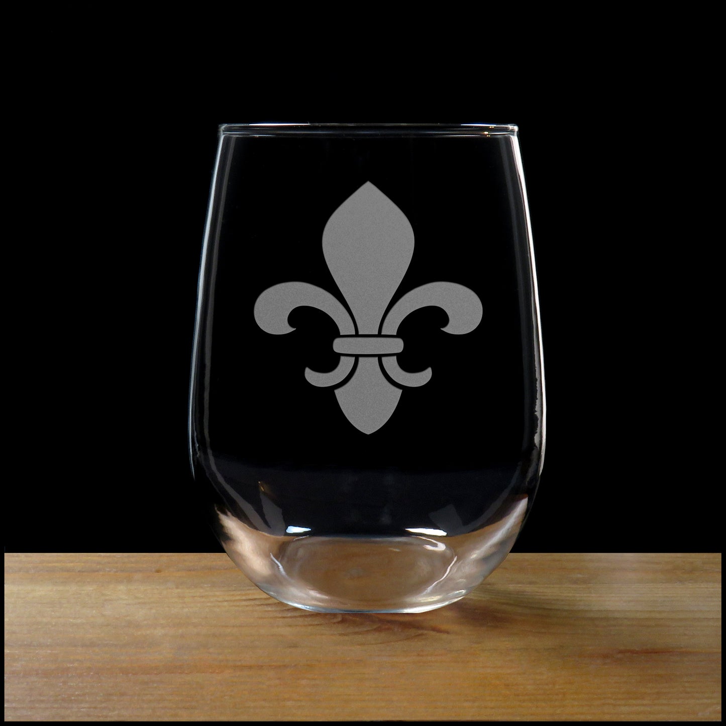 A clear stemless wine glass with a gray fleur-de-lis symbol etched on the front, resting on a wooden surface against a solid black background.