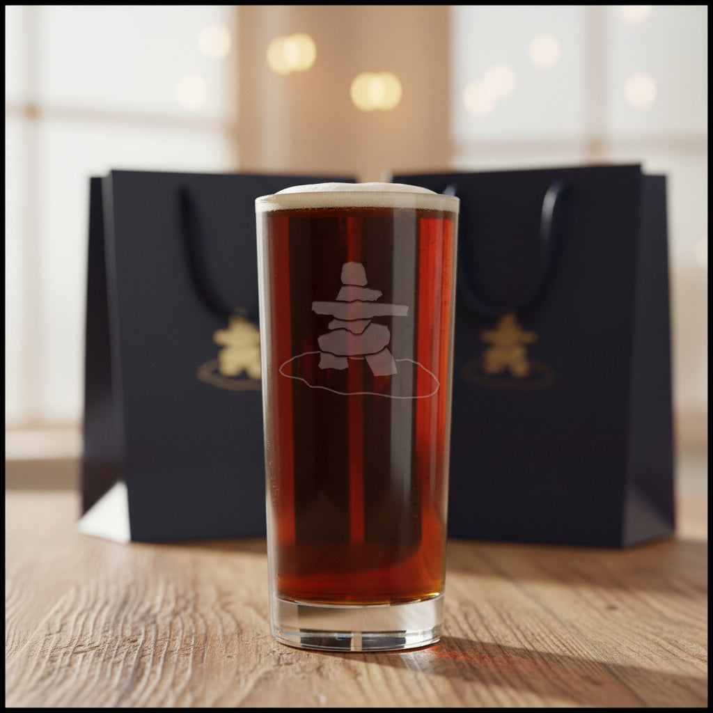 A clear pint glass filled with dark beer, featuring a frosted white etched design of a stacked stone Inuksuk on a gray textured background.