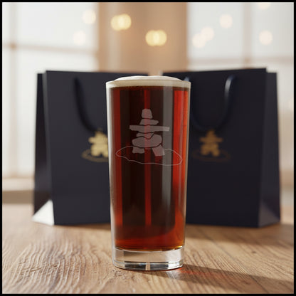 A clear pint glass filled with dark beer, featuring a frosted white etched design of a stacked stone Inuksuk on a gray textured background.