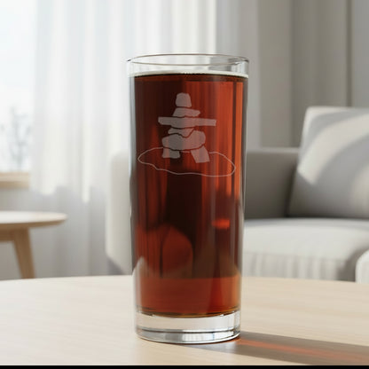 A clear pint glass filled with dark beer, featuring a frosted etched design of a Inuksuk 