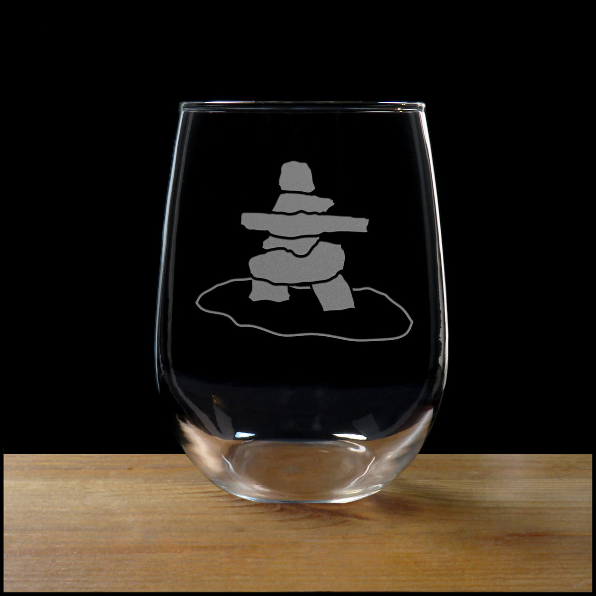 A clear stemless wine glass with a gray etched Inuit harpoon symbol, shown on a wooden surface against a solid black background.