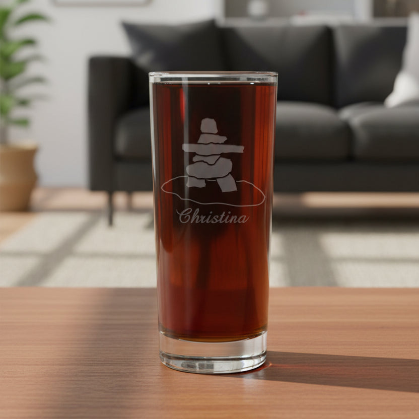 A clear pint glass filled with dark beer, featuring an etched inukluk symbol and the name 'Christina' on the front, set against a textured gray background.
