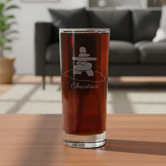 A clear pint glass filled with dark beer, featuring an etched inukluk symbol and the name 'Christina' on the front, set against a textured gray background.