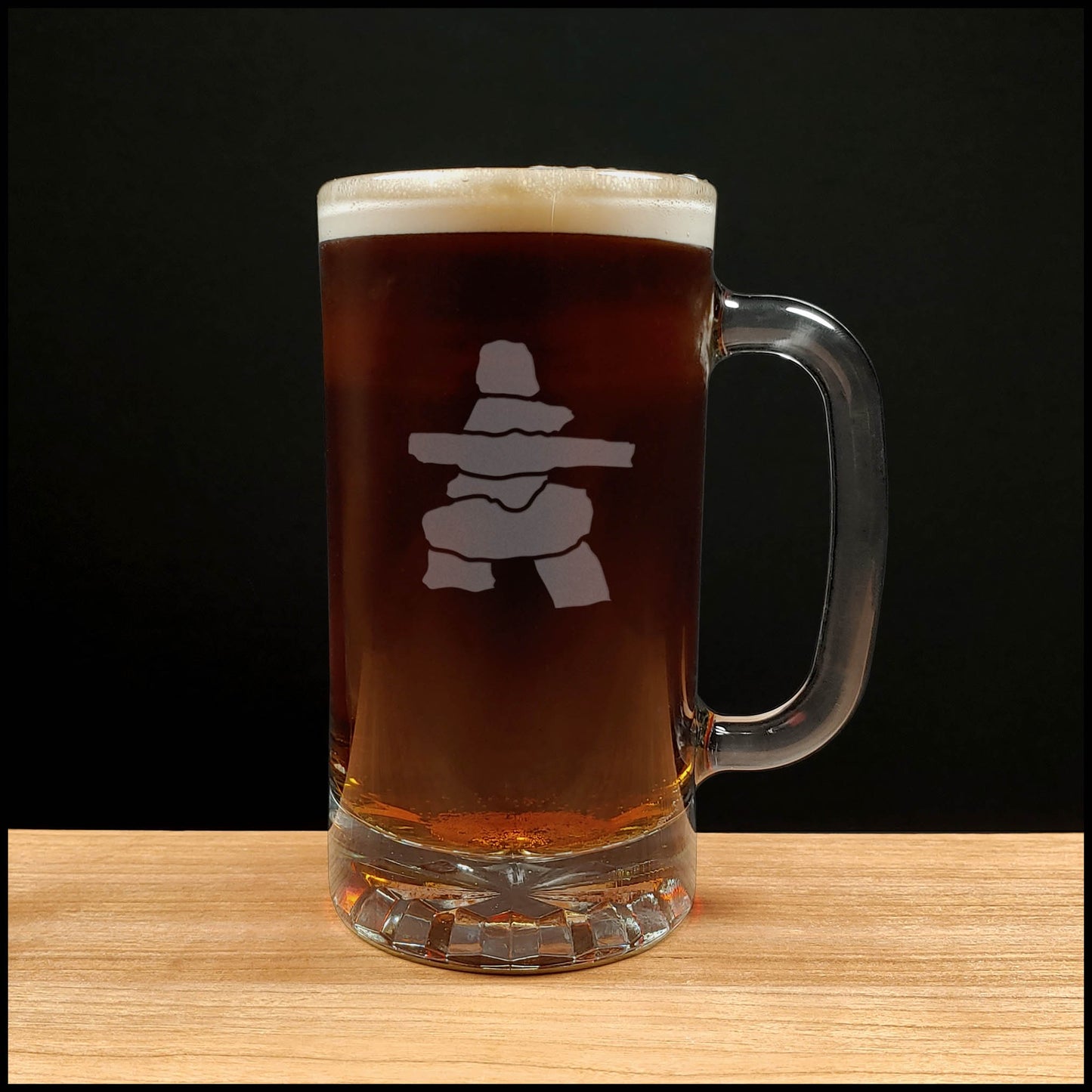A clear glass beer mug filled with dark amber beer and topped with white foam, featuring a Inuksuk design on the front, set on a light wood surface against a black background.