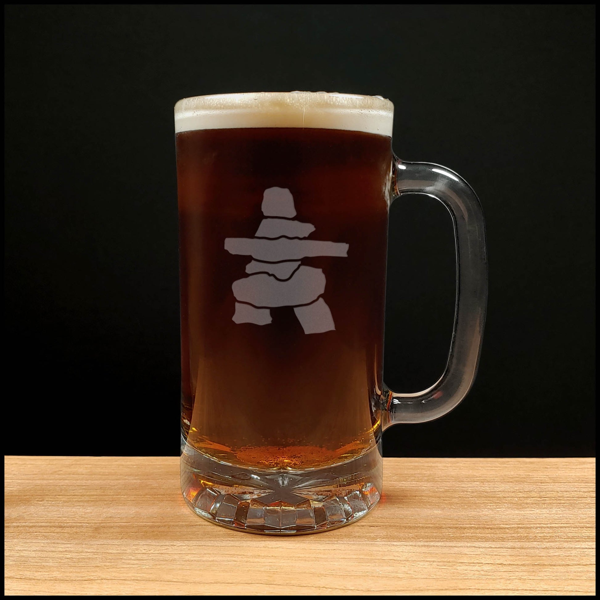 A clear glass beer mug filled with dark amber beer and topped with white foam, featuring a Inuksuk design on the front, set on a light wood surface against a black background.