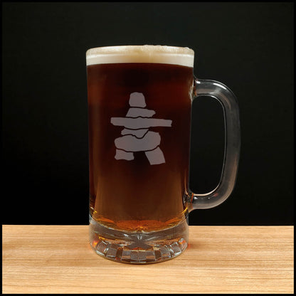 A clear glass beer mug filled with dark amber beer and topped with white foam, featuring a Inuksuk design on the front, set on a light wood surface against a black background.