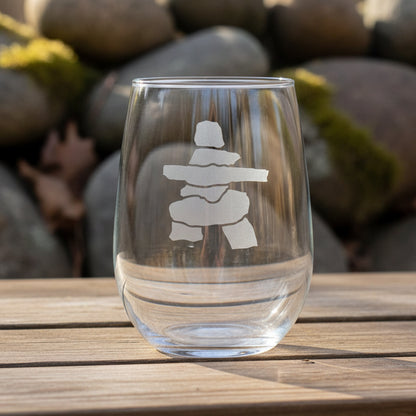 Deep sand etched 17oz stemless wine glass featuring an Inuksuk design, displayed on a wooden outdoor table with a softly blurred natural stone background. The frosted engraving stands out clearly against the transparent glass.