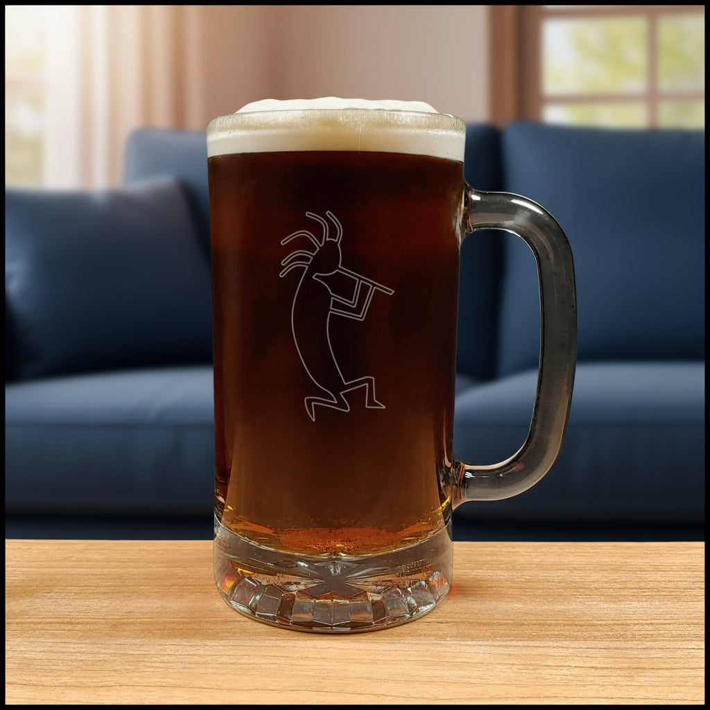 A clear glass beer mug with a handle, filled with dark amber beer and topped with white foam, featuring a etched kokopelli figure. The mug is placed on a light wood surface with a blue couch in the background.