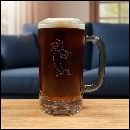 A clear glass beer mug with a handle, filled with dark amber beer and topped with white foam, featuring a etched kokopelli figure. The mug is placed on a light wood surface with a blue couch in the background.