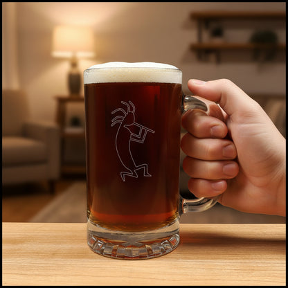 A clear glass beer mug with a handle, filled with dark amber beer and topped with white foam, featuring a etched kokopelli figure. The mug is placed on a light wood surface with a living room in the background.