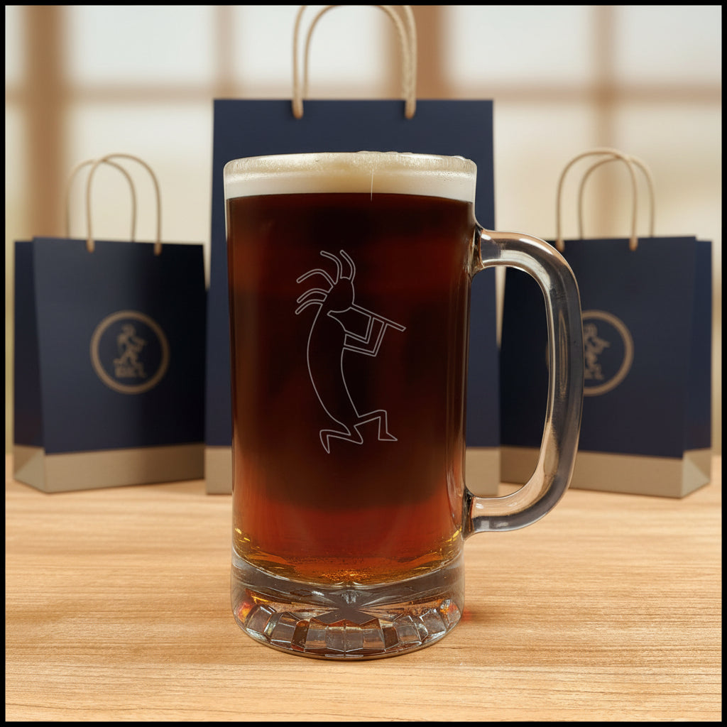 A clear glass beer mug with a handle, filled with dark amber beer and topped with white foam, featuring a etched kokopelli figure. The mug is placed on a light wood surface with a gift bags in the background.