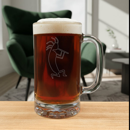A clear glass beer mug with a handle, filled with dark amber beer and topped with white foam, featuring a etched kokopelli figure. The mug is placed on a light wood surface with a living room in the background.