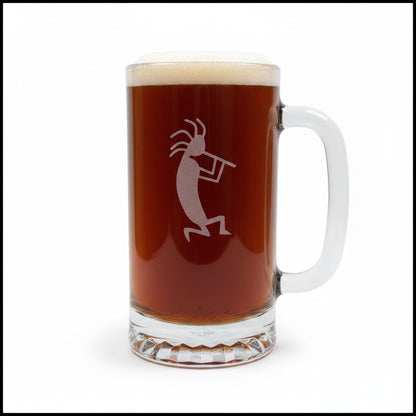 A clear glass beer mug with a handle, filled with dark amber beer and topped with white foam, featuring a etched kokopelli figure. The mug is placed on a light wood surface with a white background.
