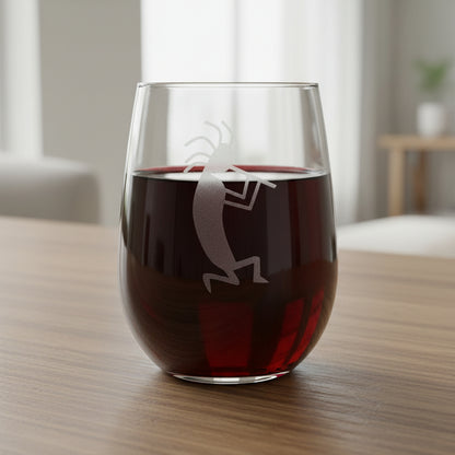 A clear stemless wine glass with a white line-art engraving of a Kokopelli dancer, resting on a wooden surface 