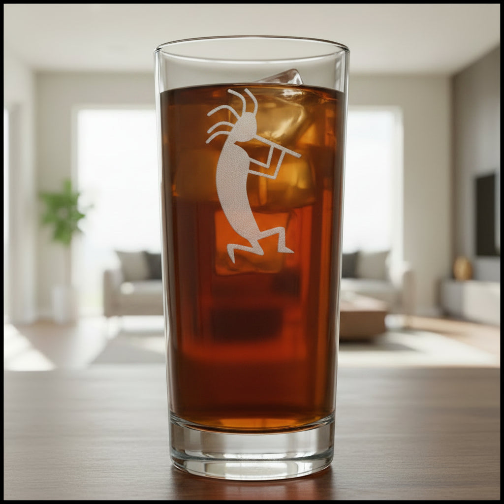 A clear highball glass filled with a dark reddish-brown liquid, featuring a white etched Kokopelli figure on the front, set against a modern living room